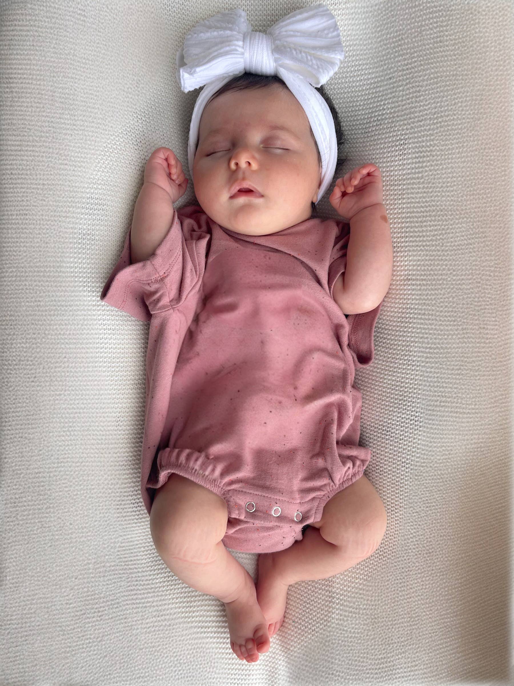 Adorable baby sleeping in mauve speckle t-shirt bubble with white bow headband, perfect for warm weather.