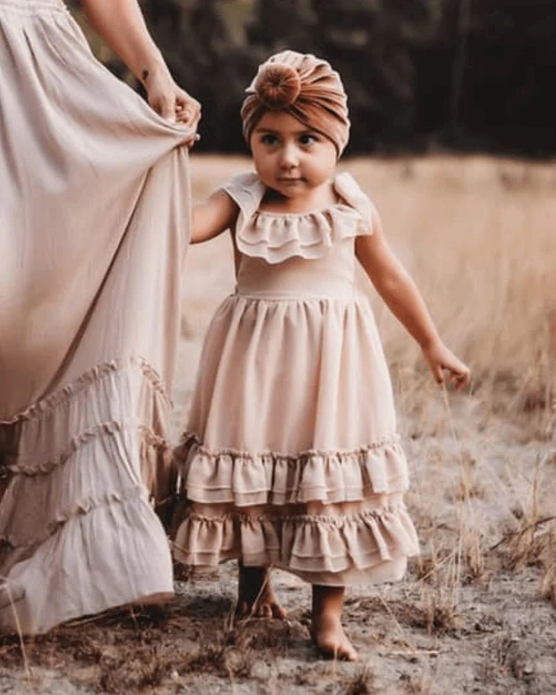 Little girl in a champagne dress with ruffles, styled with a matching headband, standing in a natural setting.