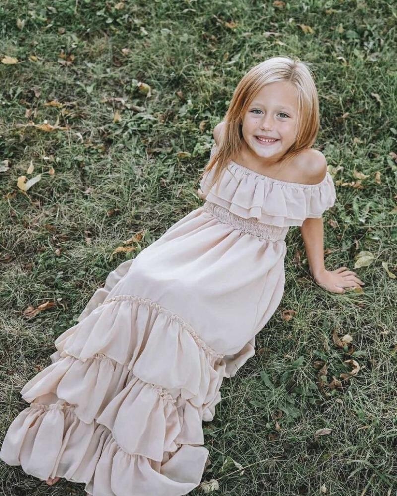 Little girl smiling in a champagne dress with ruffles, sitting on grass, wearing an off-shoulder style for special occasions.