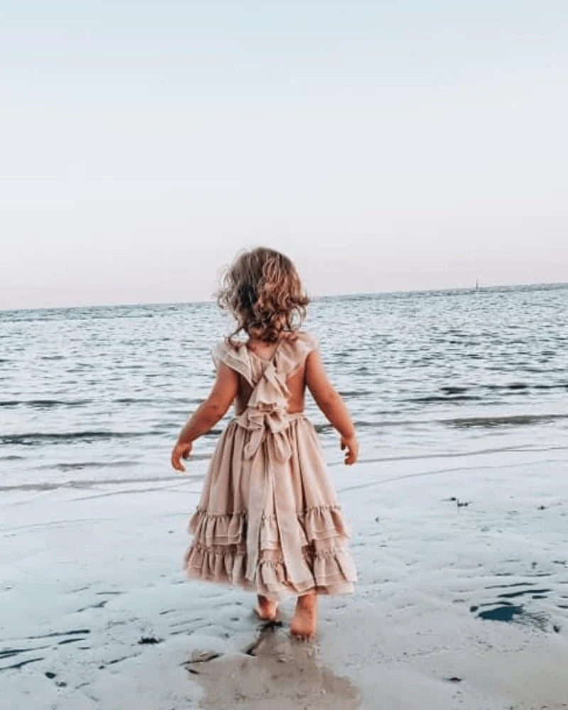 Girl wearing a champagne ruffle maxi dress walking on the beach by the water's edge.