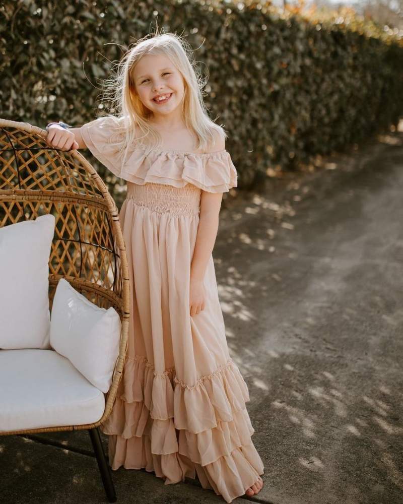 Girl in a champagne dress for girls, featuring smocking and ruffles, standing near a wicker chair.