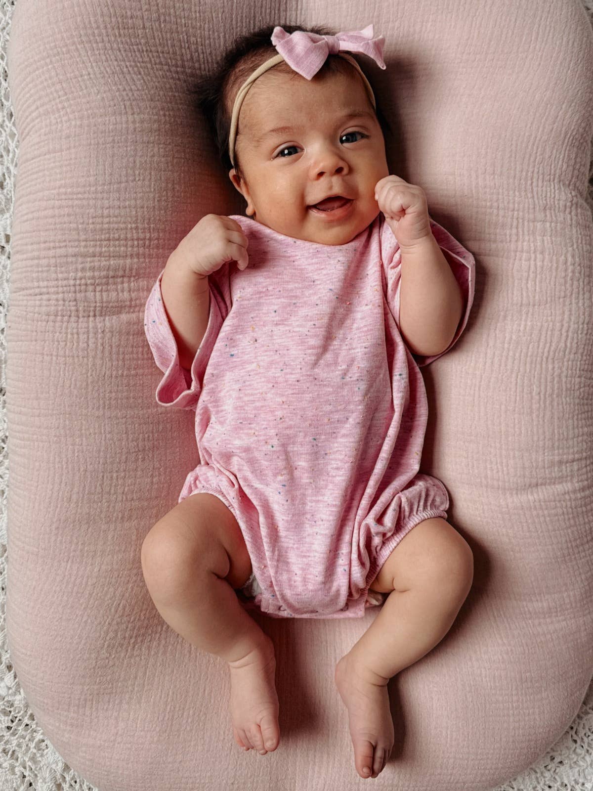 Smiling baby girl in a pink speckle t-shirt bubble laying on a soft pillow, wearing a matching headband.
