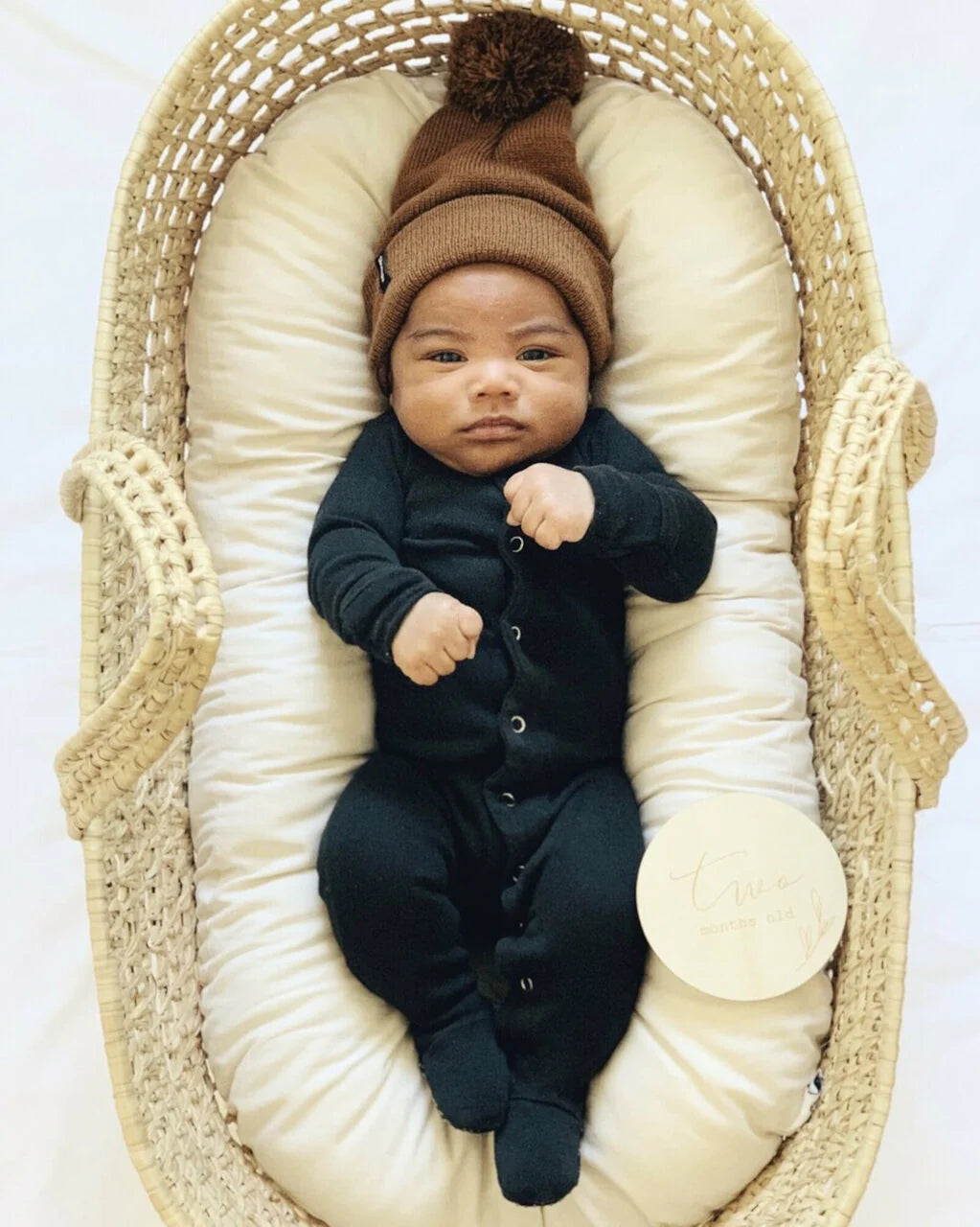 Baby wearing a black organic snap footie in a cozy basket, topped with a brown pom-pom hat.