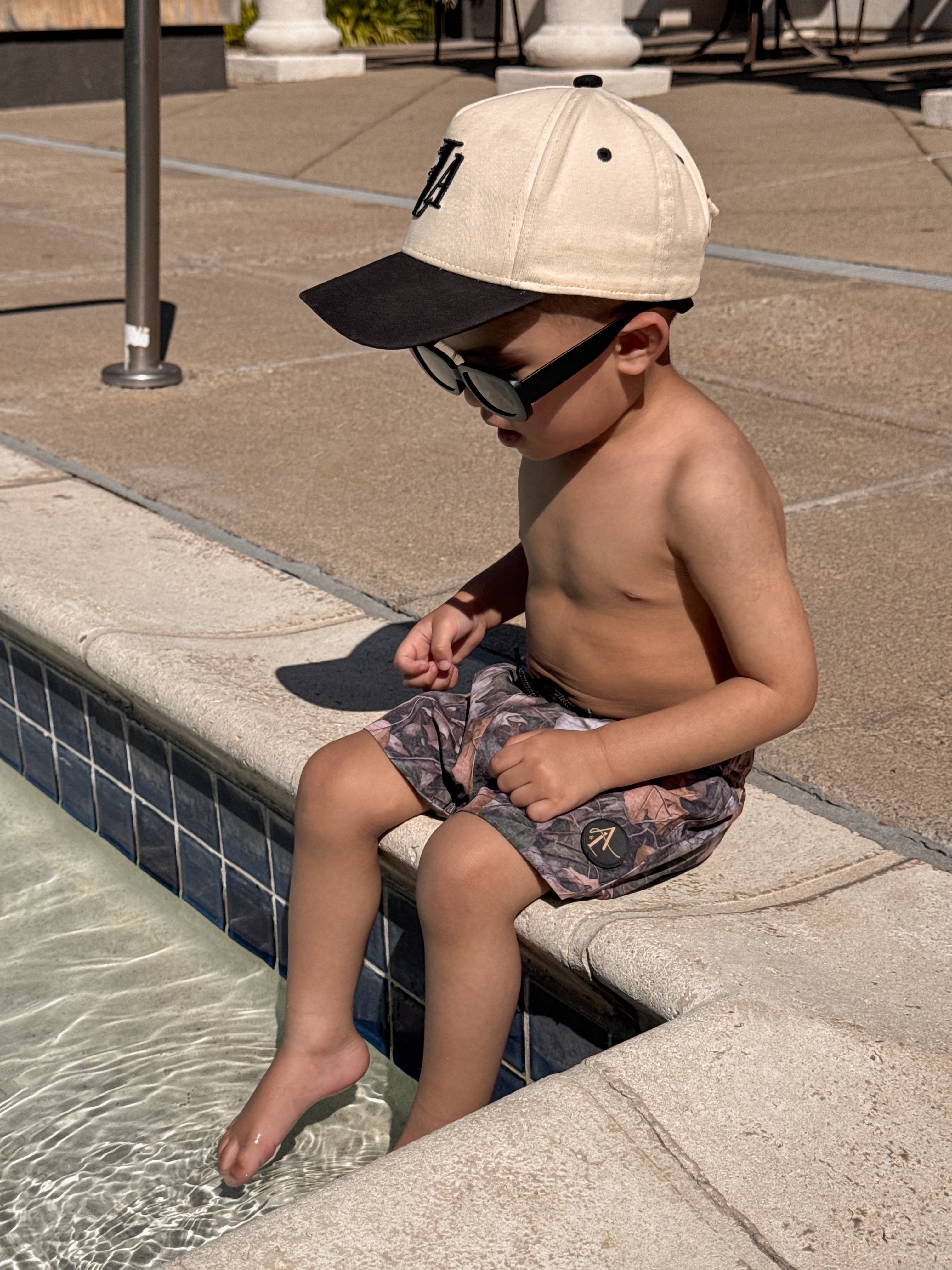 Toddler wearing camo swim shorts and a cap, sitting by the pool, enjoying a summer day.