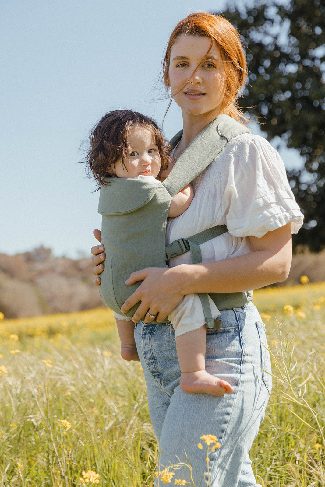 Mother carrying baby in dusky sage green Acadian Aerial Carrier in a field of flowers.